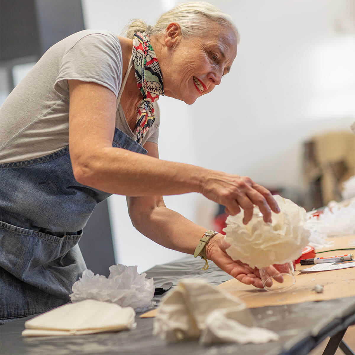 Una signora anziana sorridente, con capelli grigi raccolti e foulard colorato, lavora concentrata a un progetto di artigianato. Indossa un grembiule di jeans e tiene un tessuto bianco sopra un bicchiere; sul tavolo utensili.