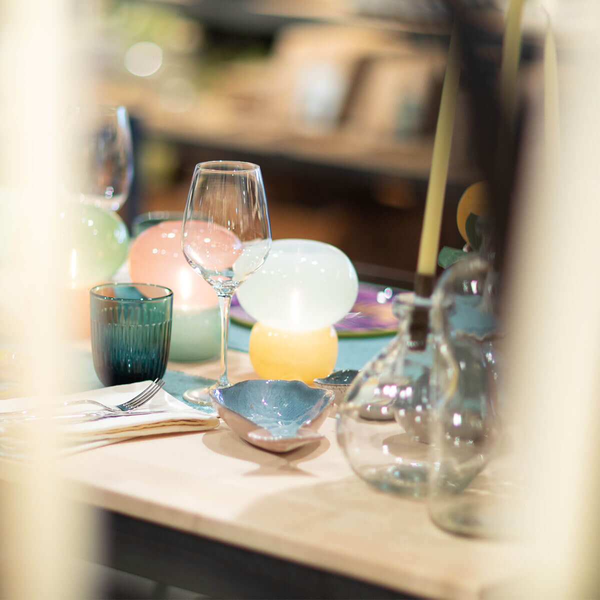 An elegantly laid table featuring an empty wine glass, a ribbed turquoise drinking glass and a small blue shell-shaped bowl. Behind them, candles in pastel shades (pink, green, white) are glowing. Warm light, a cosy, blurred background.