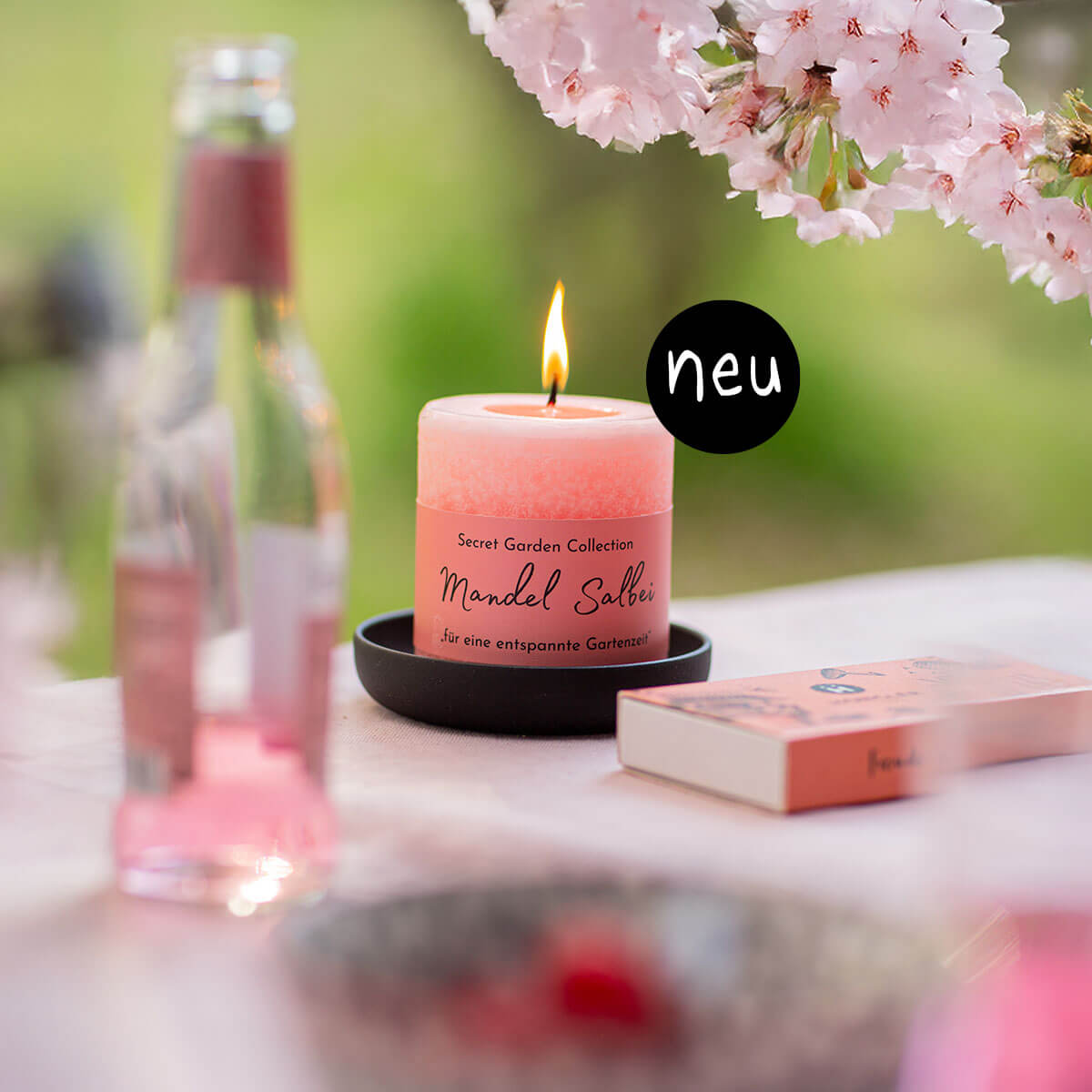 Brennende lachsfarbene Duftkerze „Mandel Salbei“ auf dunklem Teller. Im verschwommenen Hintergrund rosa Kirschblüten, grüner Garten, rosa Getränkeflasche und orange Box. Auf Kerze schwarzer Kreis „neu“.