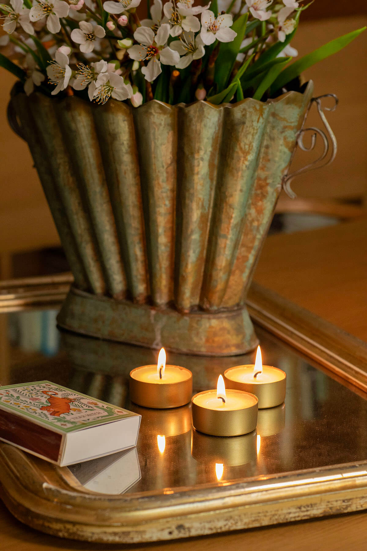 Auf einem goldenen Tablett stehen ein blumengeschmückter Topf, drei brennende Teelichter und eine Streichholzschachtel mit Tiger-Motiv. Die Kerzen spiegeln sich auf der Oberfläche des Tabletts. Das Licht erzeugt eine warme und gemütliche Atmosphäre.