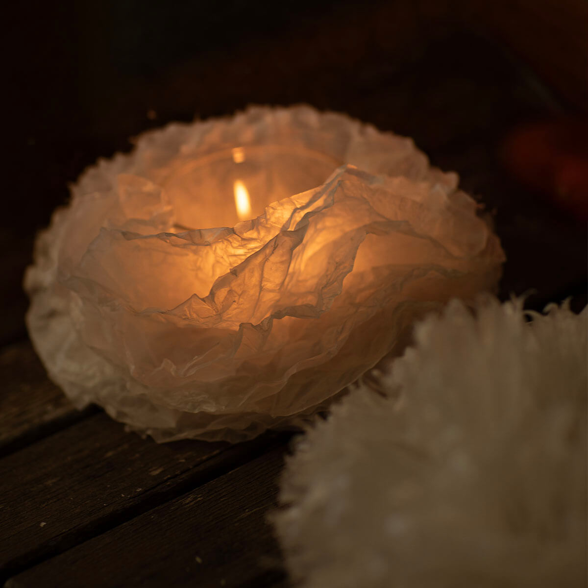 Zoom: portacandela rotondo in carta bianca piegata, simile a un fiore stilizzato o a una nuvola. Al centro arde una piccola fiamma che illumina delicatamente l'interno della carta di un colore giallo-arancio. Su tavole di legno scuro.