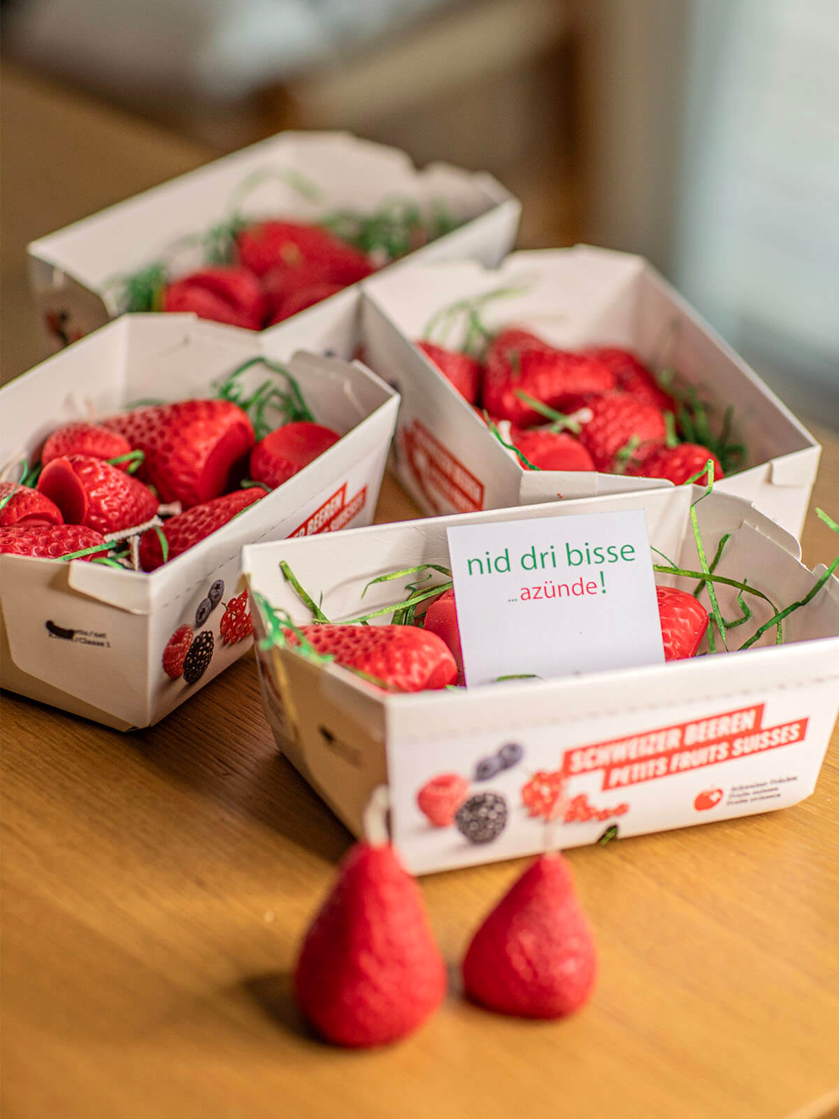 On a wooden table are four small white cardboard boxes containing strawberries and green paper strips. On top of one of the boxes is a small card with the text ‘Don’t bite into them... light them up!’. In the foreground lie two more red strawberries.