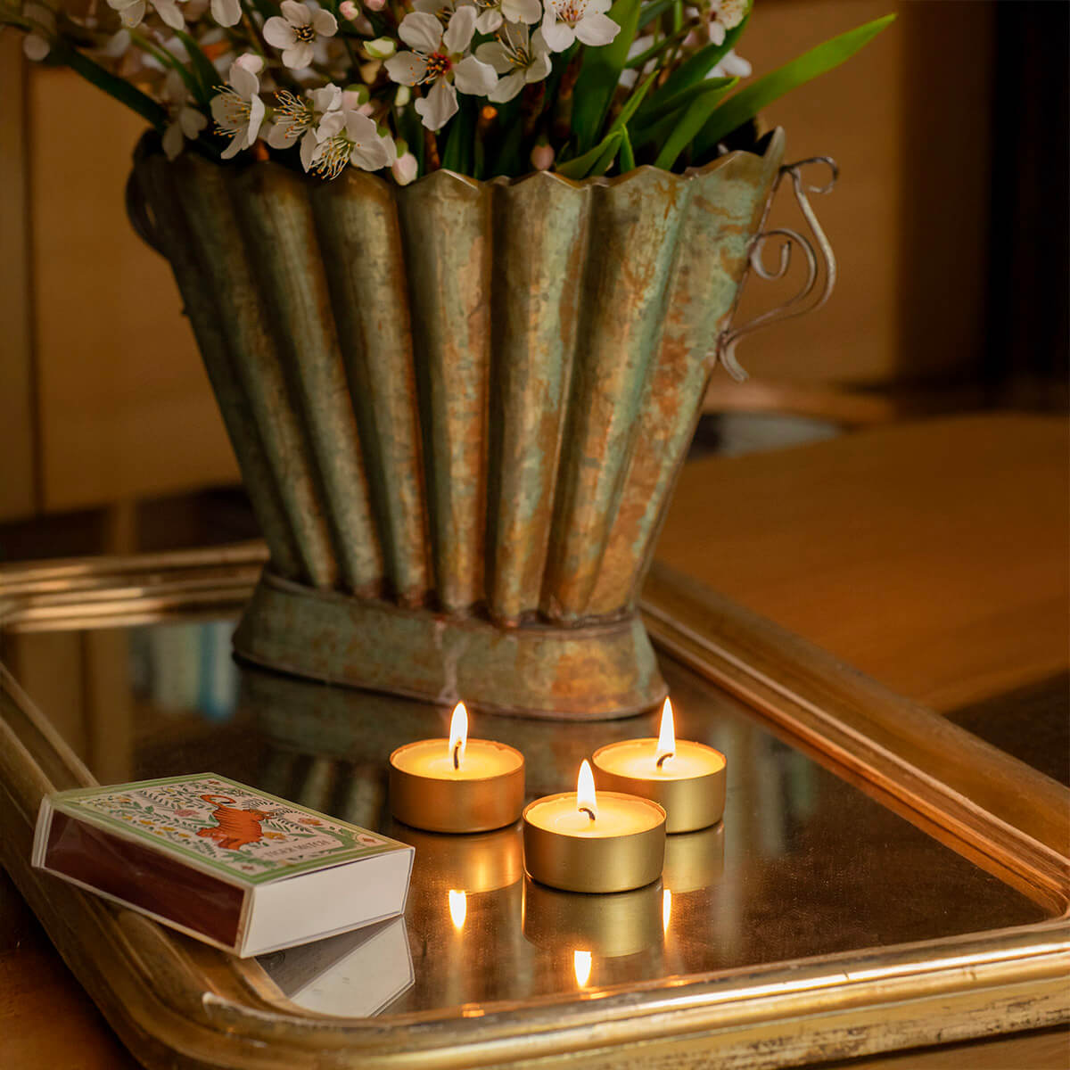 Vase vintage en métal strié patiné, rempli de fleurs de cerisier blanches, sur un plateau miroir à bordure dorée. Trois bougies chauffe-plat allumées dans des bougeoirs dorés projettent flammes et reflets. Boîte d’allumettes.