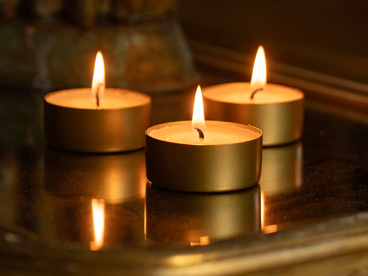 Drei Teelichter stehen auf einer spiegelnden Oberfläche. Die Flammen der Kerzen werfen ein warmes Licht und spiegeln sich auf der Oberfläche, was die Szene zusätzlich erhellt. Die goldenen Kerzenhalter tragen zu der warmen, beruhigenden Atmosphäre bei.