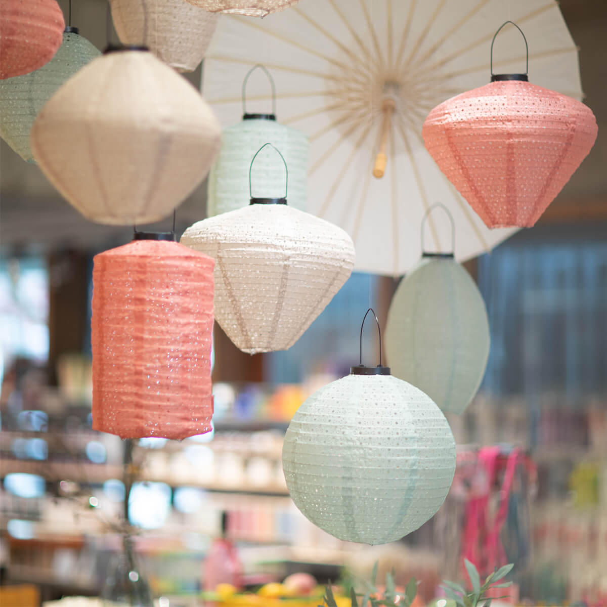 Hanging paper lanterns in pastel shades such as pale pink, mint green and cream white float through the room. They range from cylindrical to diamond-shaped to spherical and shimmer gently. In the background, a light-coloured paper umbrella.