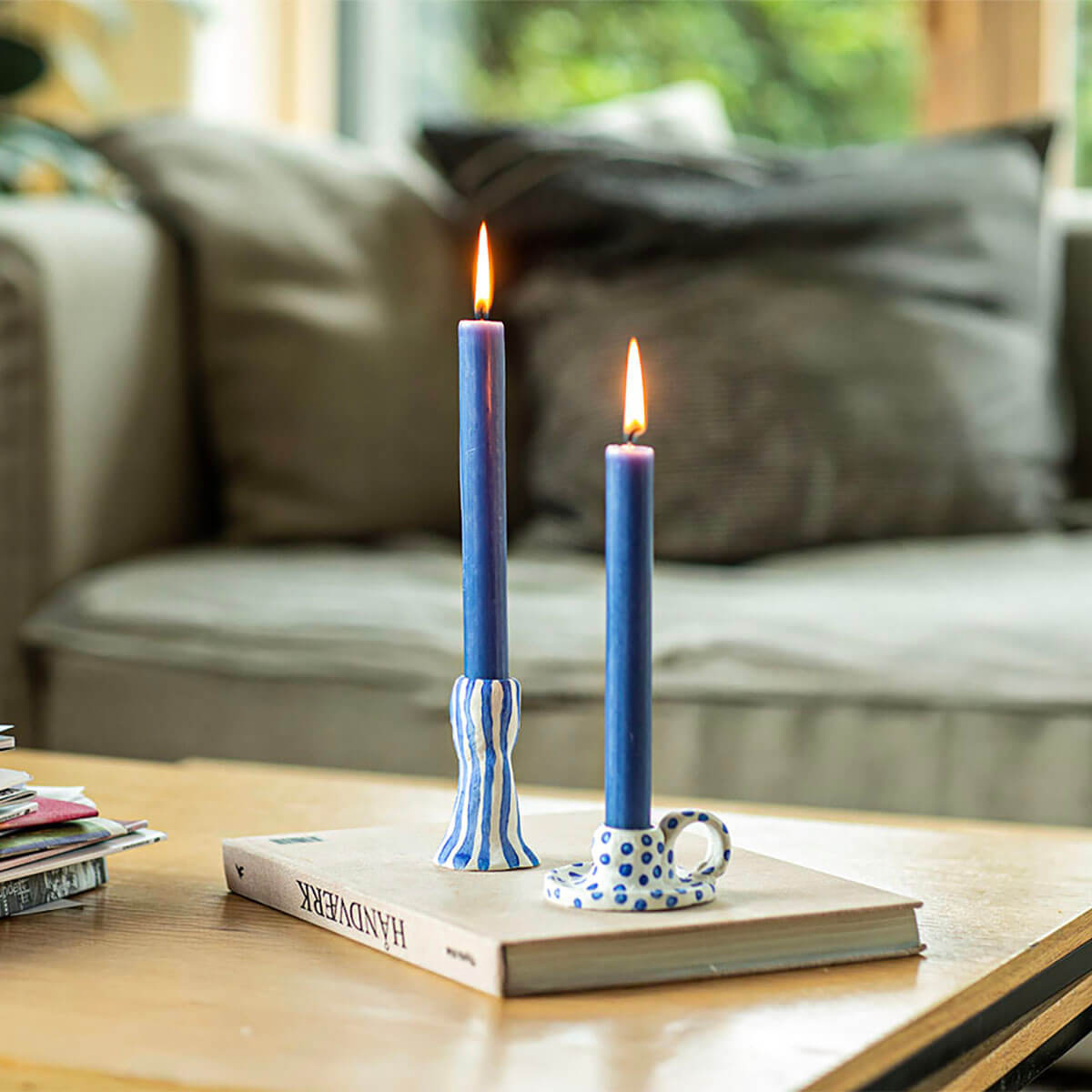 Due candele blu accese sono posizionate su un tavolo in legno. A sinistra in un portacandele a strisce bianche e blu, a destra in uno bianco con pois blu e un piccolo manico. Sotto, un libro intitolato «HANDVÆRK». Sullo sfondo, un divano e una finestra.