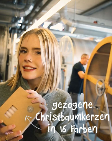 Eine junge Frau präsentiert eine Schachtel Banner Christbaumkerzen. Im Hintergrund ein Mann an einer Anlage. Die Kerzen sind in einer braunen Schachtel mit kleinem Sichtfenster verpackt. Die Frau lächelt und trägt einen grauen Pullover.