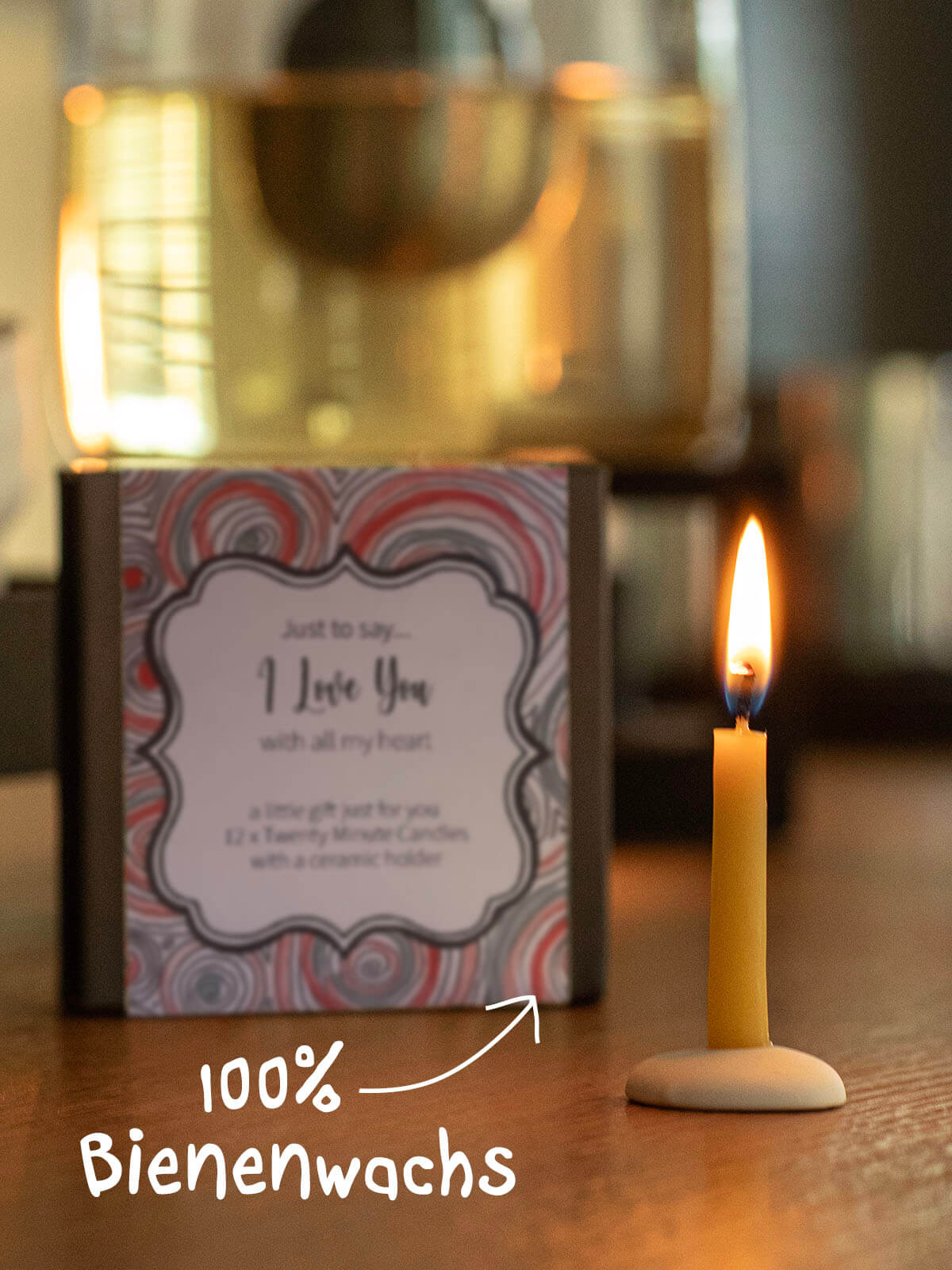 A yellow beeswax candle stands in a small ceramic holder. Next to it is a gift box with a label reading ‘I Love You’. In the background, a golden jug or vase stands on a wooden surface, blurred into the background.