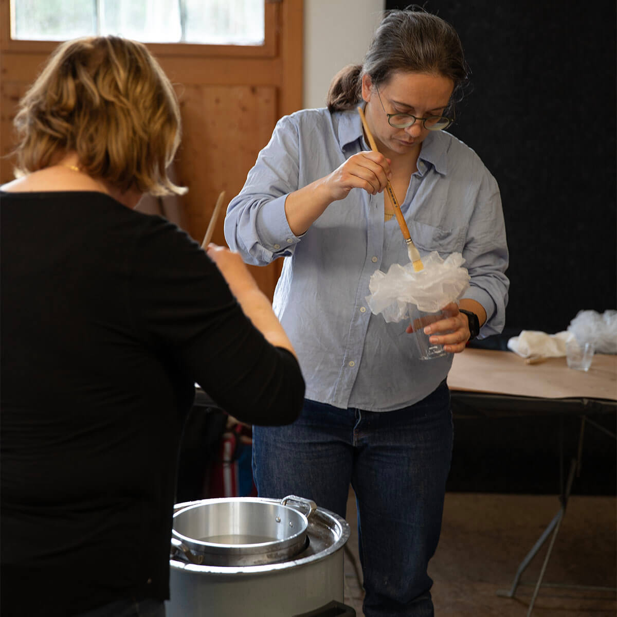 Una donna con gli occhiali e una camicia blu lavora concentrata in un atelier a un progetto artigianale, in cui con un pennello dispone dei materiali bianchi e sgualciti in un vaso di vetro. Un'altra persona è in piedi a sinistra e lavora a un vaso.