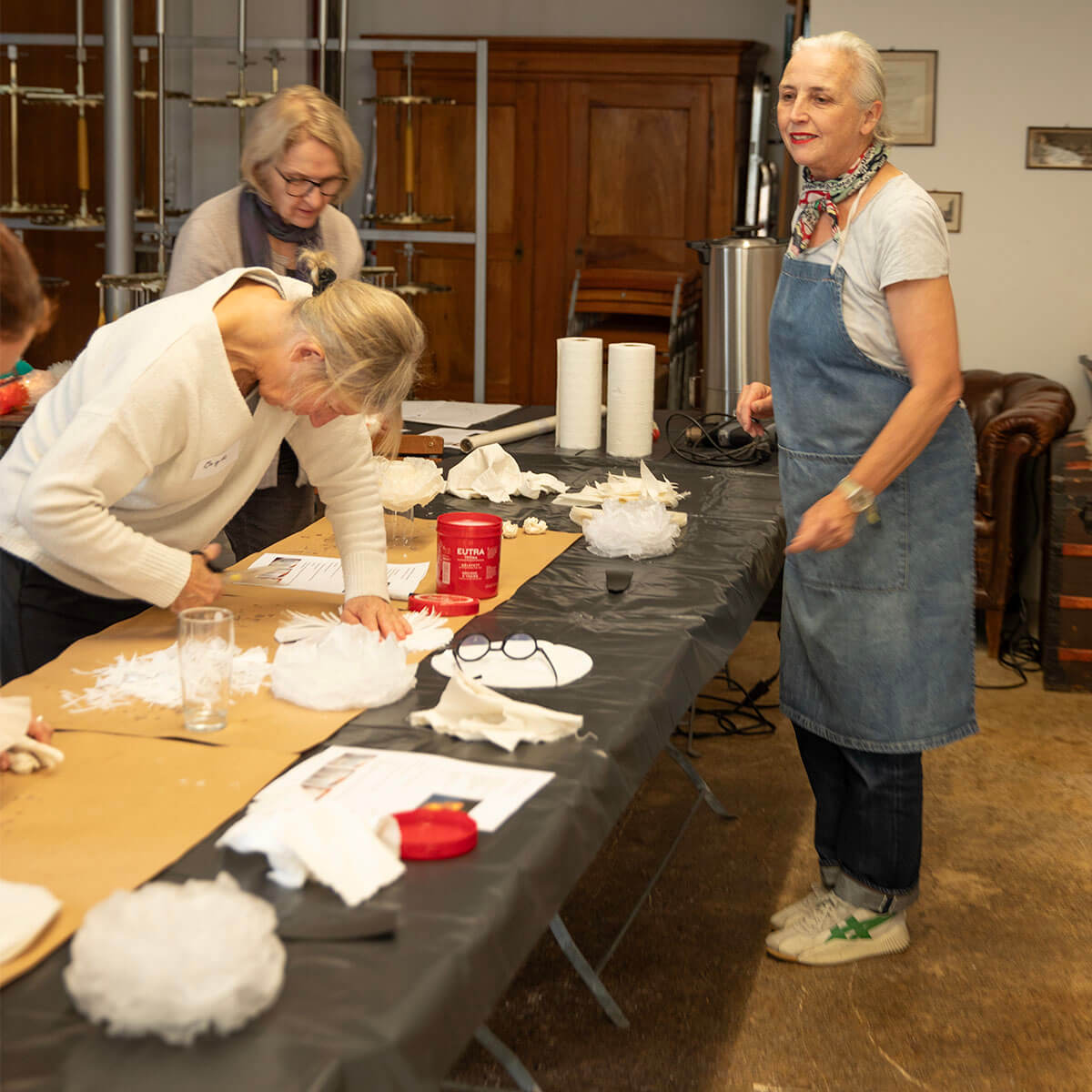 Tre donne in un workshop di bricolage. Una con un maglione bianco taglia materiale bianco per decorazioni al tavolo. Accanto, una donna sorridente con salopette di jeans e sciarpa colorata. Sul tavolo forbici, carta e un contenitore rosso.