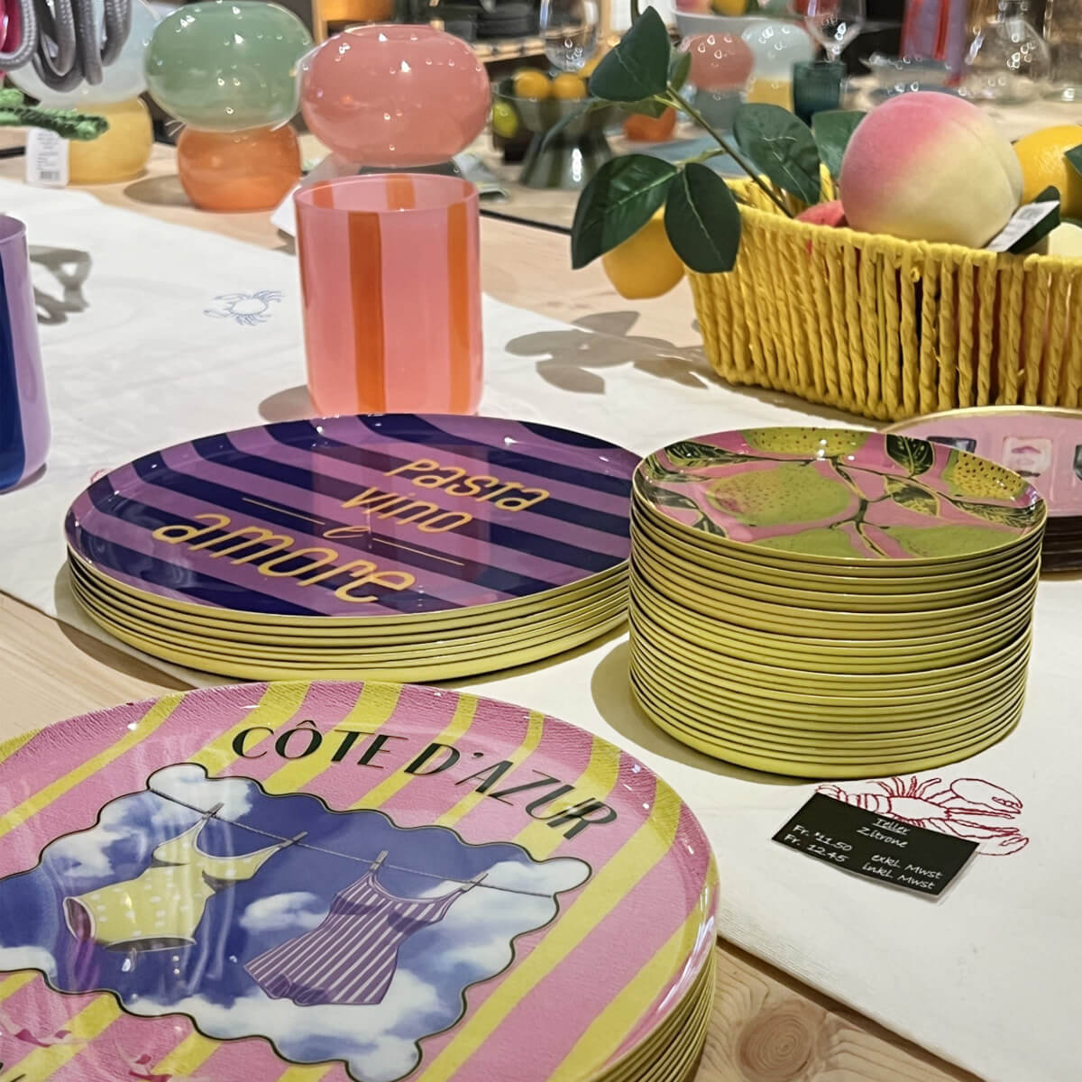 Colourful crockery and decorations on a table. Stacked plates: purple-striped with ‘pasta vino amore’, pink ones with ‘CÔTE D'AZUR’ and beachwear, small plates with a lemon pattern. Glass jars and a yellow fruit basket in the background.