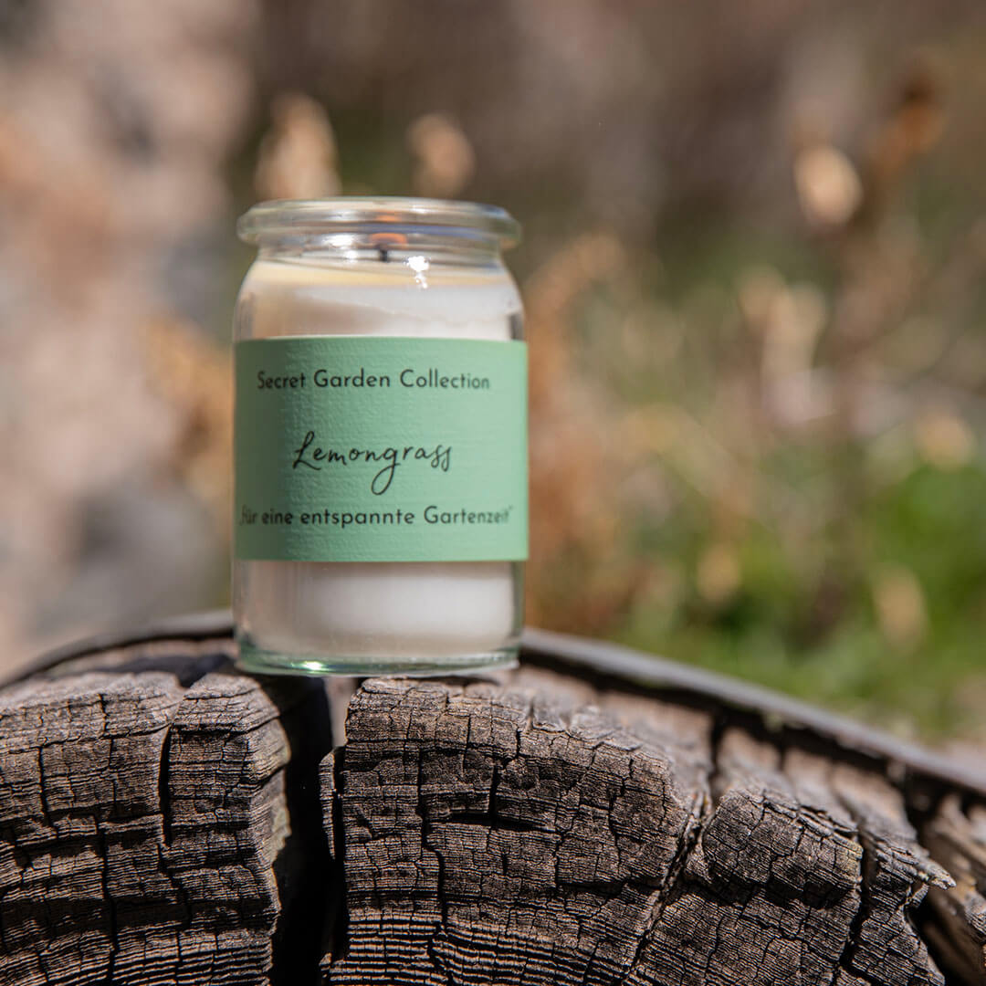 Ein zylindrisches Glasgefäß mit einer weißen Duftkerze steht auf einem alten, rissigen Holzstamm. Auf dem hellgrünen Etikett steht 'Secret Garden Collection Lemongrass für entspannte Gartenzeit'. Hintergrund verschwommen.