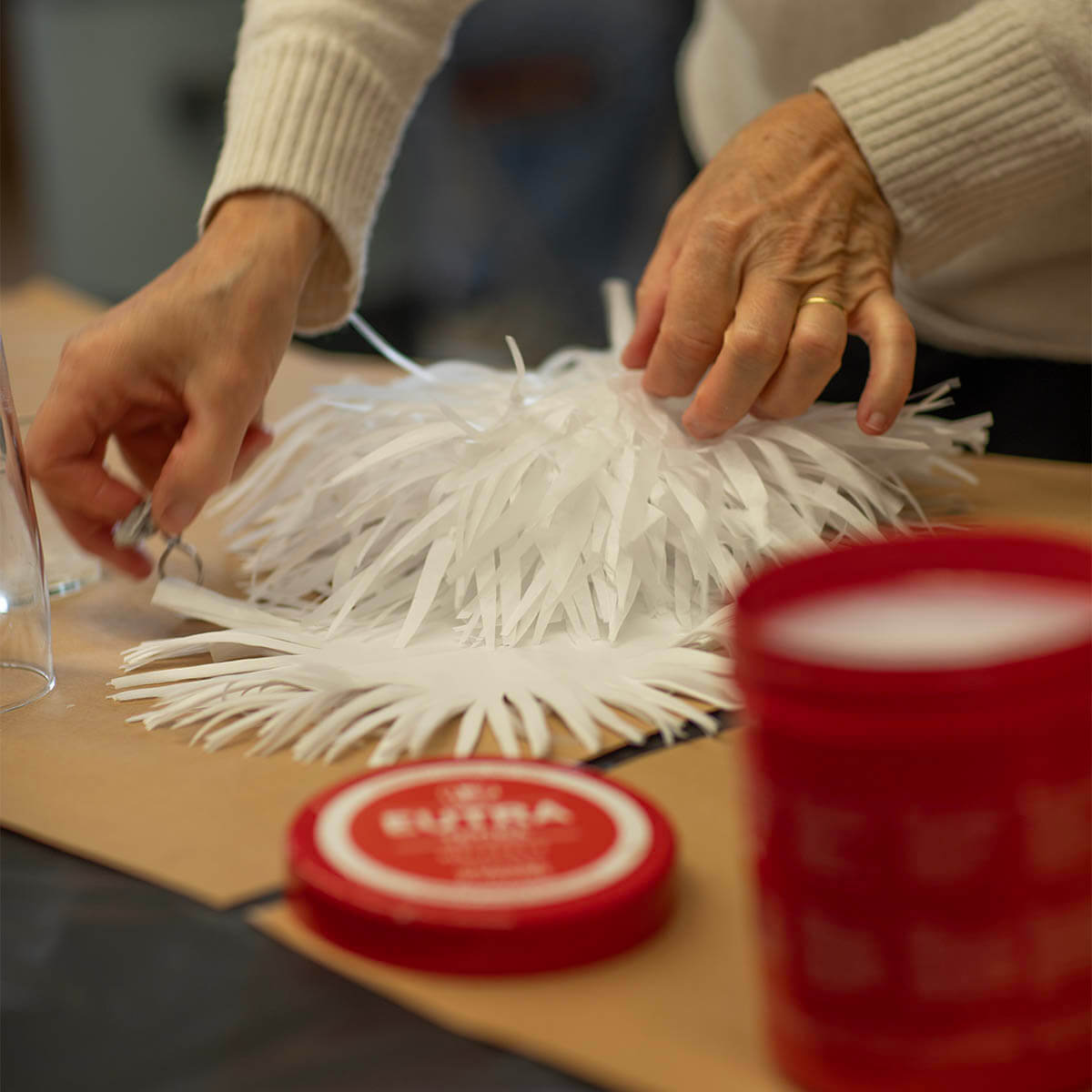 Una persona con un maglione chiaro a maglia taglia sottili strisce di carta bianca per creare una decorazione soffice. Mani con forbici e carta; ne risulta un oggetto vaporoso a forma di stella. In primo piano, sfocato, un contenitore rosso.