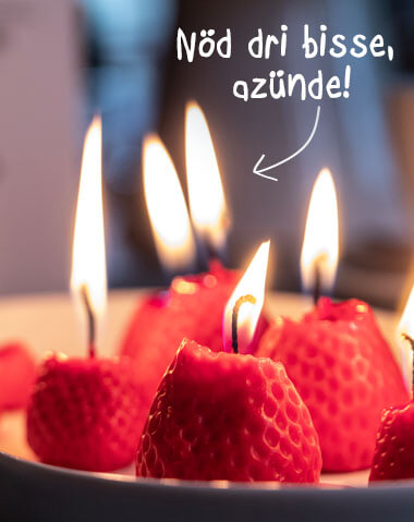 Close-up of strawberry-shaped candles with lit wicks, arranged on a plate. Above the candles, written in white lettering, is the phrase: "Don’t bite them, light them!" An arrow points to one of the candles. The candles are strawberry red in colour.