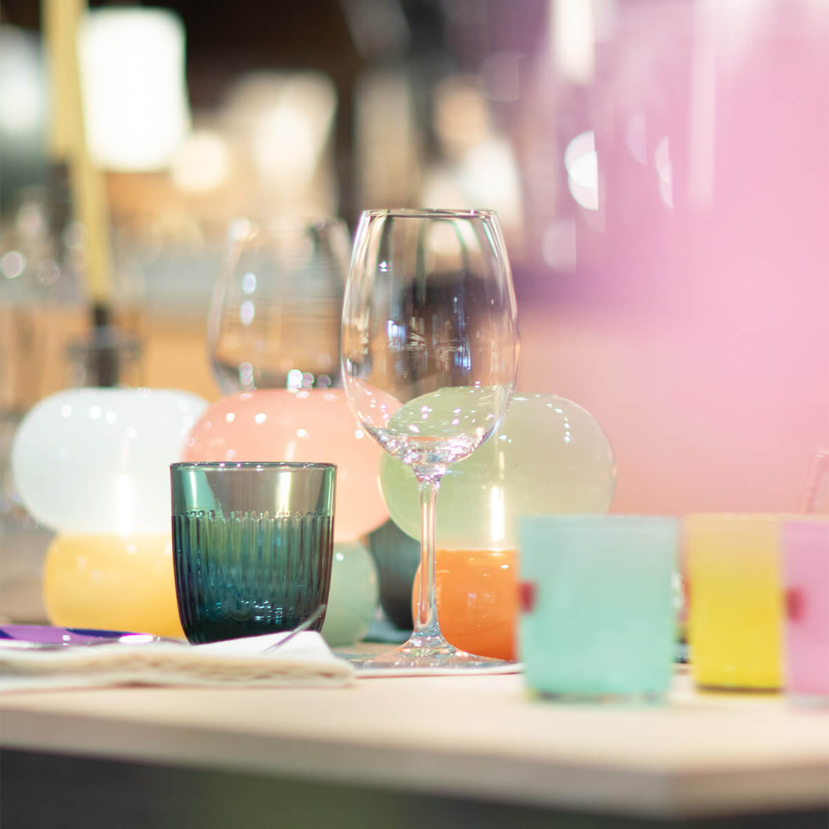 Table setting featuring a clear wine glass and a dark blue tumbler in the foreground. Behind them, spherical decorative elements in pastel shades such as peach, light green and white catch the light. Colourful glasses in the background.