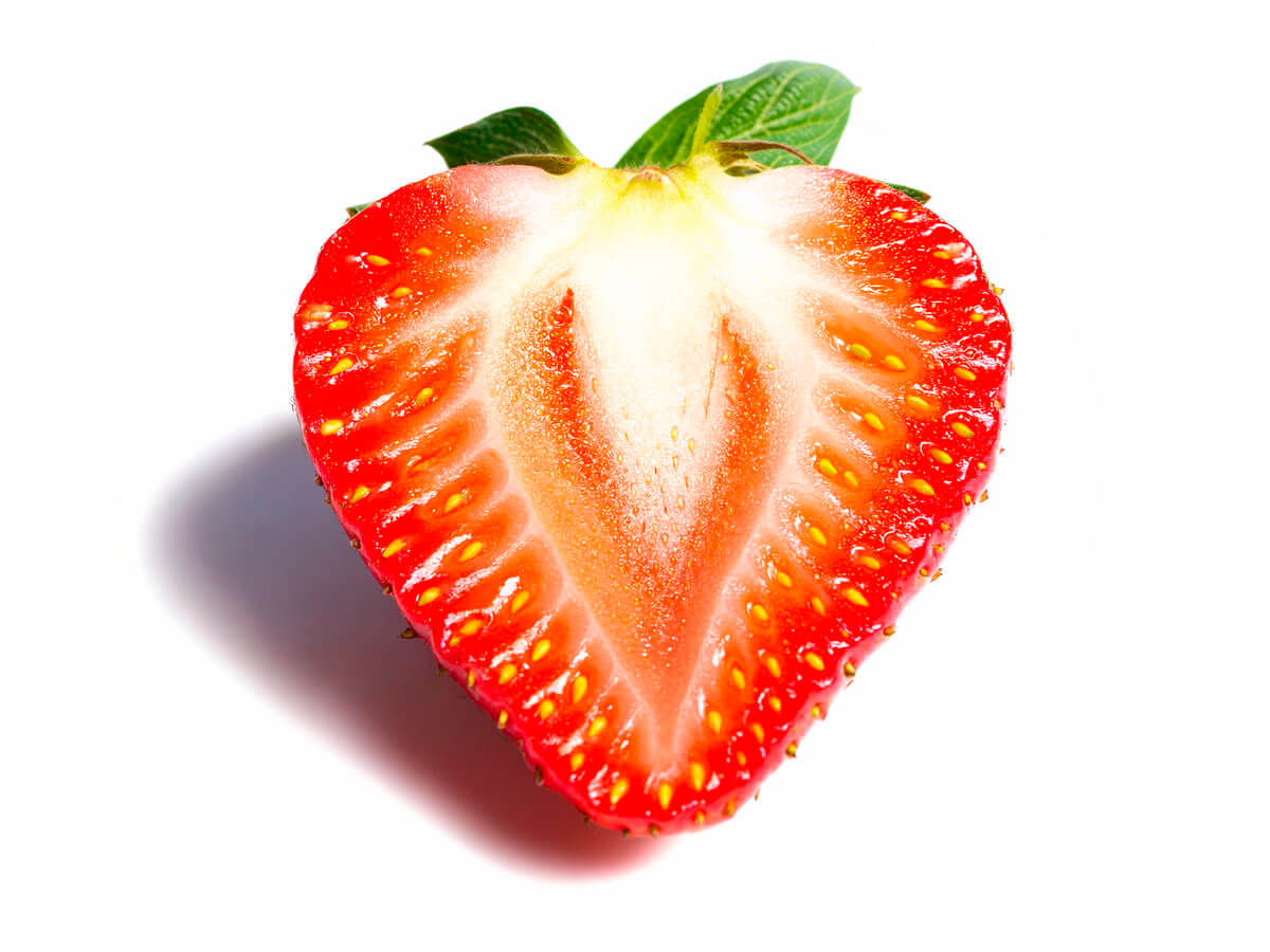A strawberry has been cut in half lengthways. The flesh is white to pale red and has small yellow seeds. Green leaves can be seen at the top. The background is white and a slight shadow can be seen on the left.