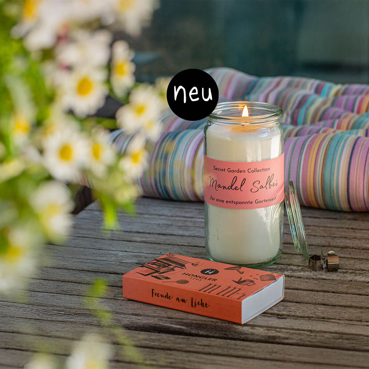 Neu: Brennende "Mandel Salbei" Duftkerze im Glas auf Holz, mit rosa Etikett "Secret Garden Collection" und "entspannte Gartenzeit". Daneben Streichholzschachtel. Im Hintergrund Gänseblümchen und buntes Kissen.
