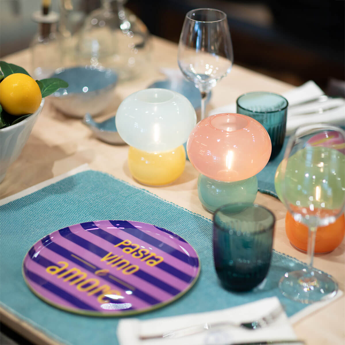 A stylishly laid dining table with a turquoise tablecloth and purple plates bearing the yellow lettering ‘Pasta Vino Amore’. Colourful mushroom-shaped candle holders in pastel yellow, pink and green, with wine glasses, mugs, lemon wedge and cutlery.