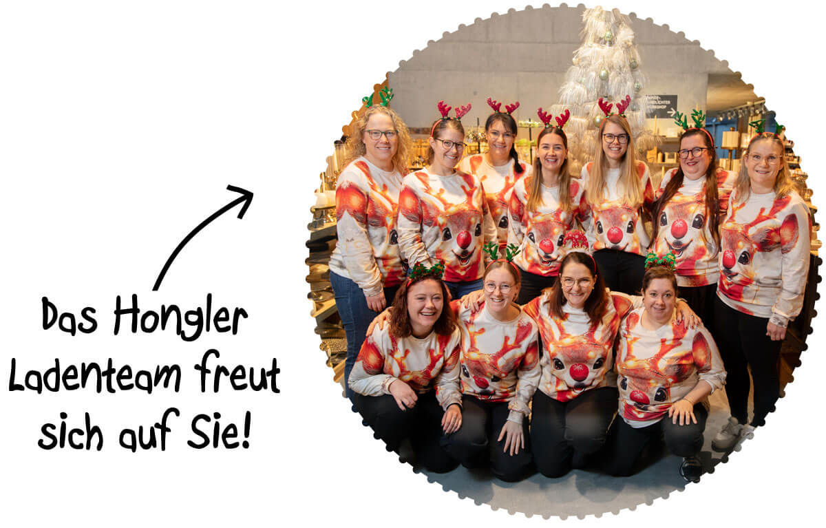 Zehn lächelnde Frauen, das Hongler Ladenteam, posieren in festlichen Rentierpullovern mit roter Nase und Rentier-Geweihen. Sie stehen und knien vor einem unscharfen Hintergrund, der eine Ladenumgebung mit einem weißen Weihnachtsbaum zeigt.