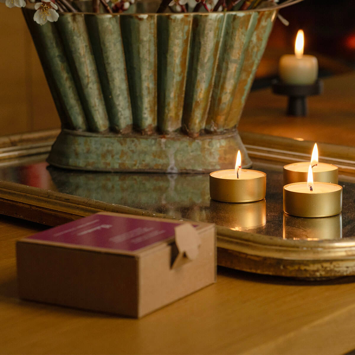 Des bougies chauffe-plat à la lueur chaleureuse sur un plateau en miroir doré. Au premier plan, une petite boîte marron au couvercle violet sur la table en bois. Derrière, un vase vert vieilli strié contient des branches fleuries blanches.