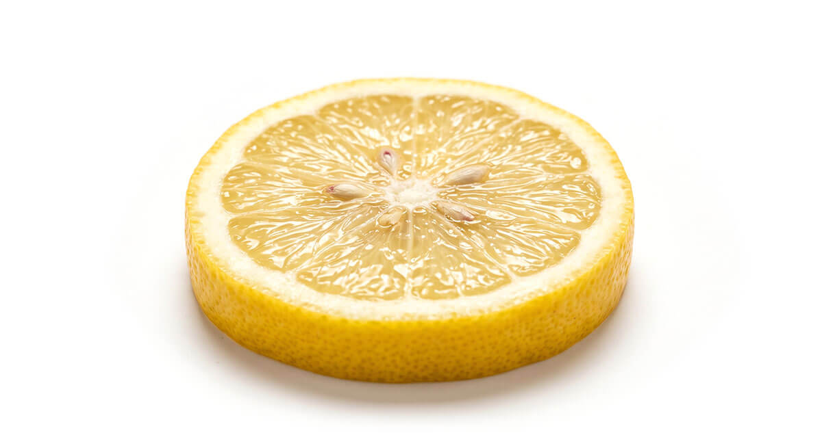 Una fetta di limone fresca e rotonda giace su uno sfondo bianco. La polpa gialla, con spicchi a raggiera e alcuni semi marrone chiaro, è chiaramente visibile, circondata da un bordo bianco e da una buccia di un giallo brillante.
