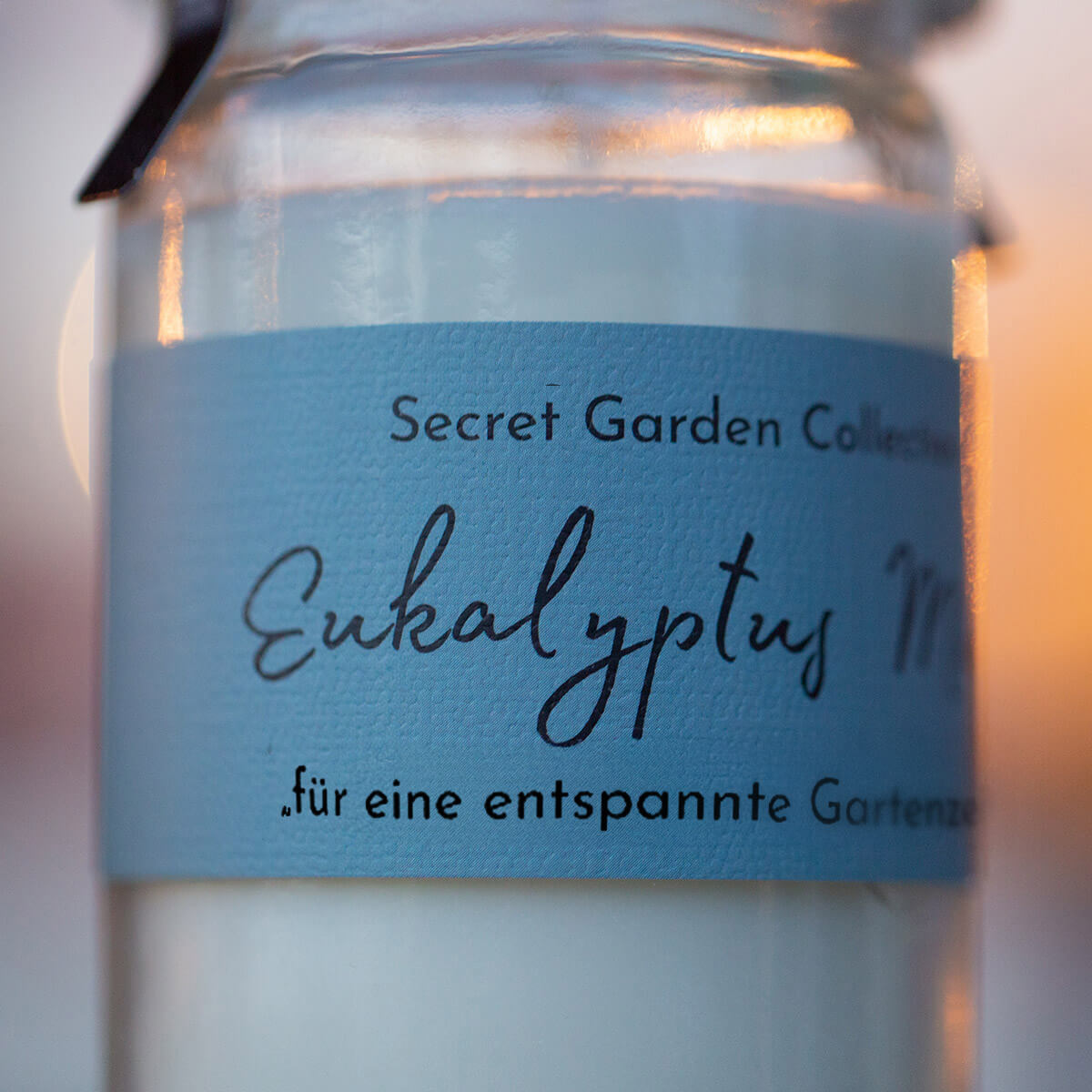 Nahaufnahme eines Glasgefäßes mit hellblauem Etikett: 'Secret Garden Collection' und groß 'Eukalyptus' in Kursivschrift. Darunter 'für eine entspannte Gartenzeit'. Enthält weiße Substanz, warmes unscharfes Licht.