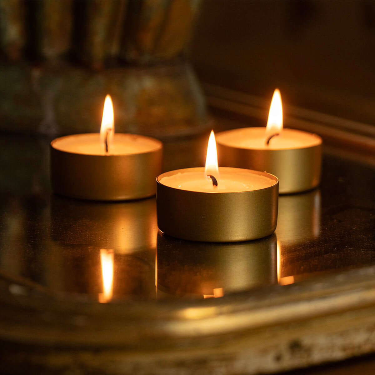 Trois bougies chauffe-plat allumées dans des supports dorés sur une surface sombre et réfléchissante; leurs flammes et reflets créent une ambiance chaleureuse, avec un fond doré flou.