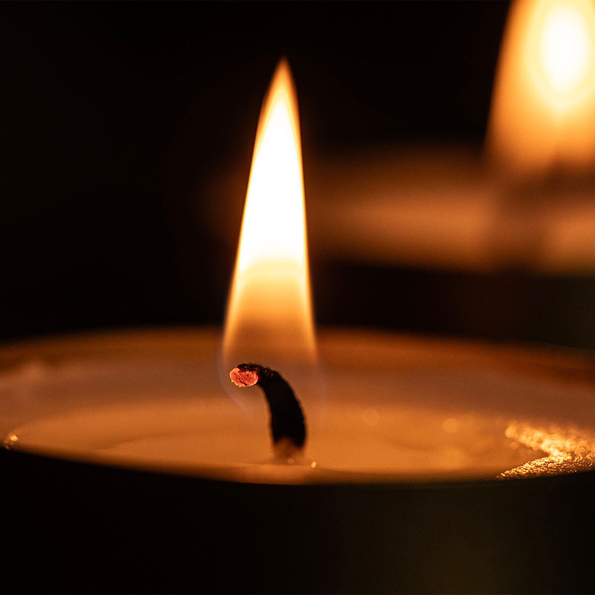 Gros plan sur une bougie allumée dans une pièce sombre. La flamme orange-jaune, nette au centre, éclaire la cire fondue dans le bougeoir. À l'arrière-plan flou, une autre bougie brille faiblement, créant une atmosphère chaleureuse.