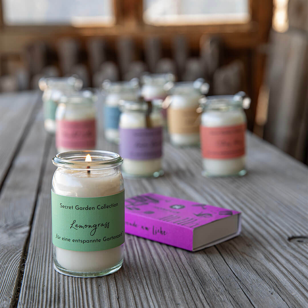 Eine brennende Kerze in klarem Glas mit grünem 'Lemongrass'-Etikett steht auf einem rustikalen Holztisch. Daneben liegt eine magenta Streichholzschachtel 'made with liebe'. Im unscharfen Hintergrund weitere Duftkerzen.