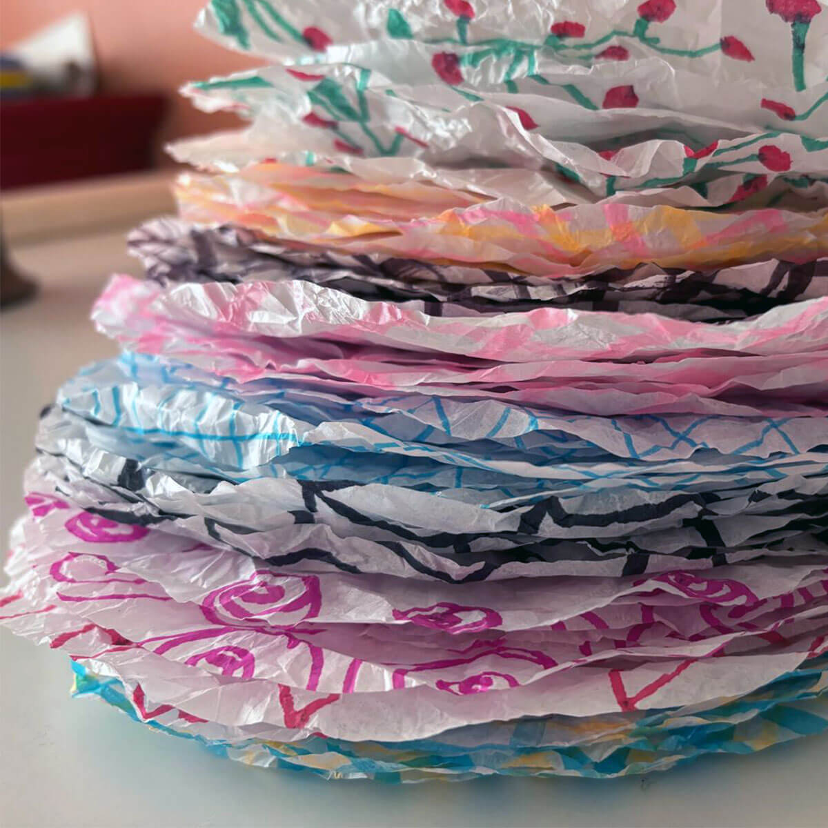 Primo piano di una pila di dischi di carta stropicciati e traslucidi. Ogni strato circolare presenta motivi colorati: bacche rosse su steli verdi, spruzzi giallo-rosa, linee nere, onde rosa e reticoli blu.