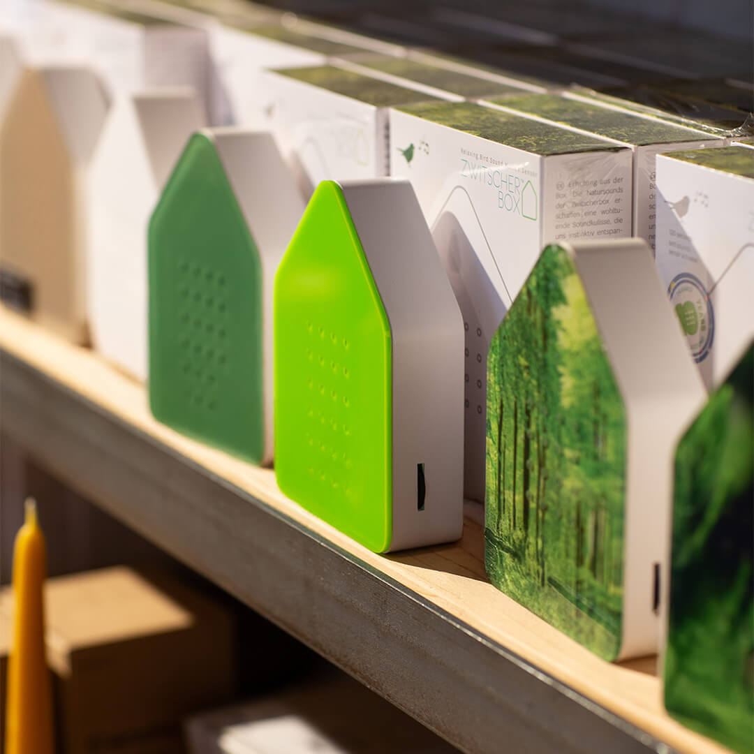 There are several house-shaped devices on a shelf. The casings are white and the gable fronts are green, light green or printed with a forest motif. A small slot can be seen on the side of each device. Some of the devices are packed in cardboard boxes.