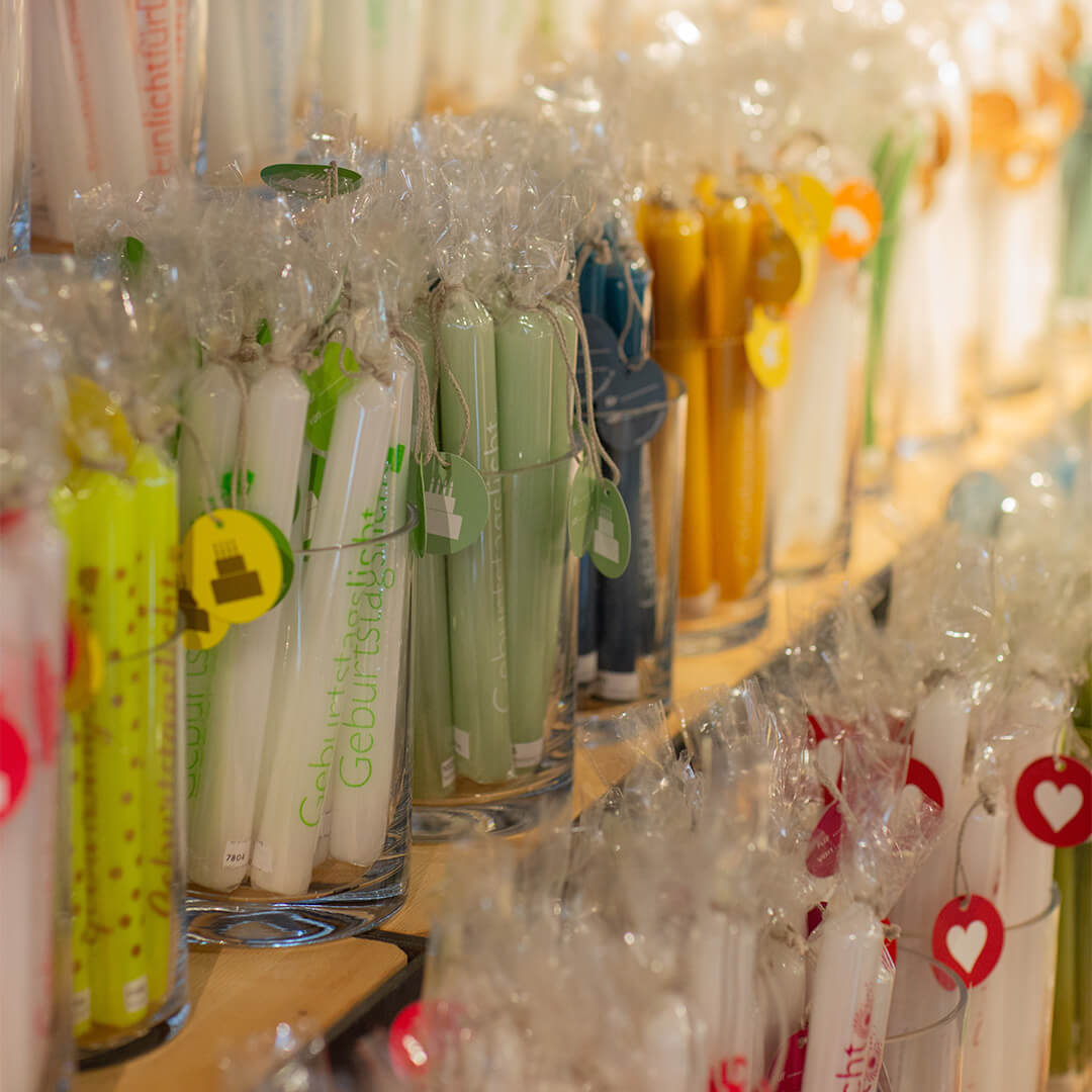Candles of various colours in glasses, wrapped in foil, are standing on a shelf. Each group of candles has a small coloured cardboard label. Some of the candles bear the words ‘birthday candle’.