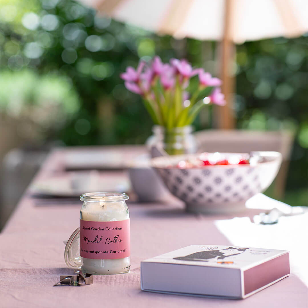 Brennende Duftkerze mit rosa Etikett „Mandel Salbei“ auf hellrosa Tischläufer. Daneben eine kleine weiße Box mit Katzenmotiv. Im Hintergrund Schale mit roten Früchten und Vase mit rosa Blumen vor Garten mit Sonnenschirm.
