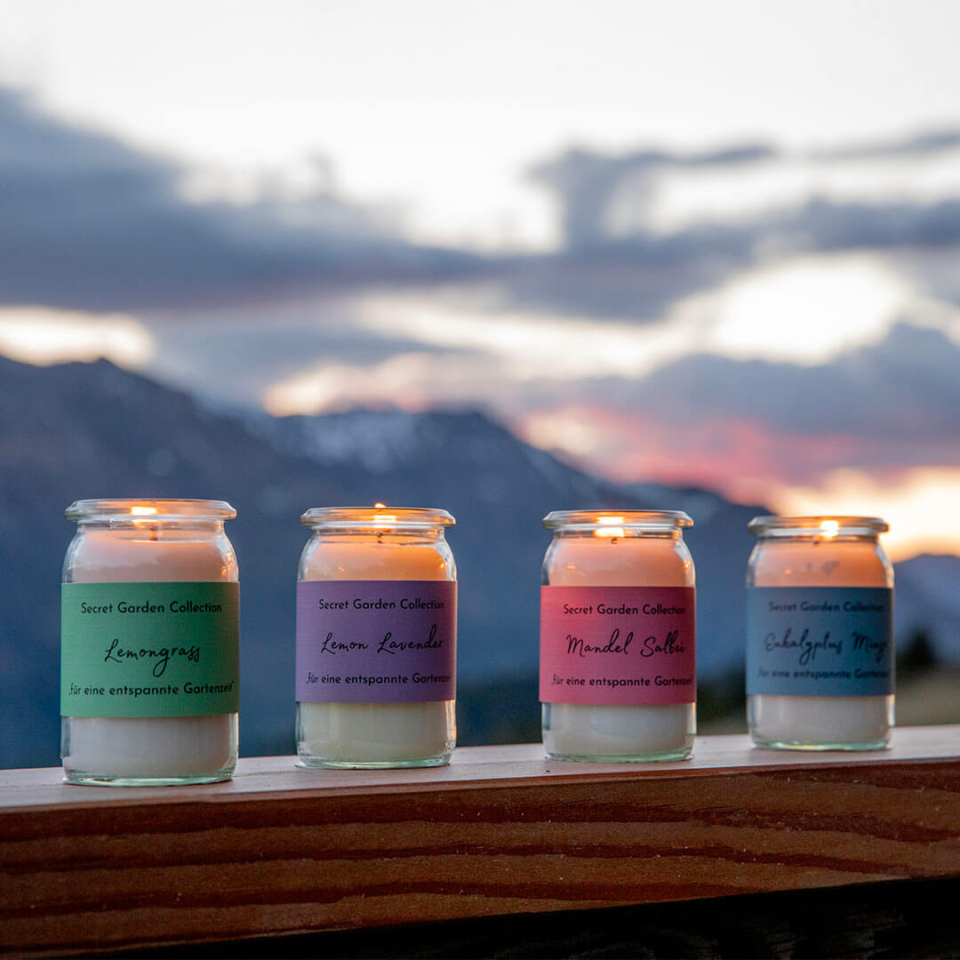 Vier brennende Duftkerzen in Glasgefäßen stehen auf dunkler Holzleiste vor Bergkulisse bei Sonnenuntergang. Etiketten: Lemongrass (grün), Lemon Lavender (violett), Mandel Salbei (rosa), Eukalyptus Minze (blau). Himmel orange-lila.