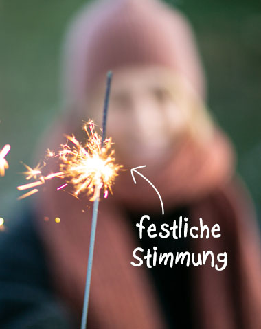 Eine Person hält eine brennende Wunderkerze in der Hand. Funken sprühen von der Spitze der Wunderkerze. Im Hintergrund ist eine weitere Person mit Mütze und Schal unscharf zu erkennen. Über die Wunderkerze ist der Text "festliche Stimmung" gelegt.